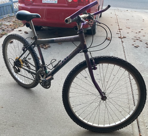 A dusty and dirty bicycle. It has purple metallic handlebar grips, a front wheel that's not pointing in the same direction as the handlebars, a yellowed dork disc, and a rusty geartrain.